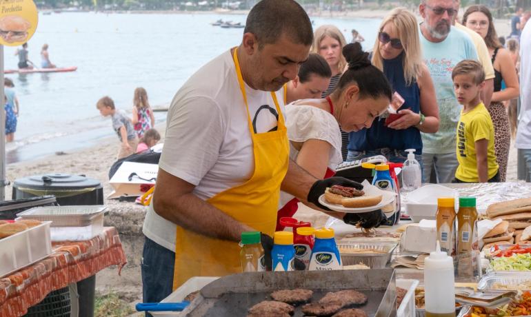 Grigliata in spiaggia, persone in fila per hamburger, atmosfera vivace e conviviale.