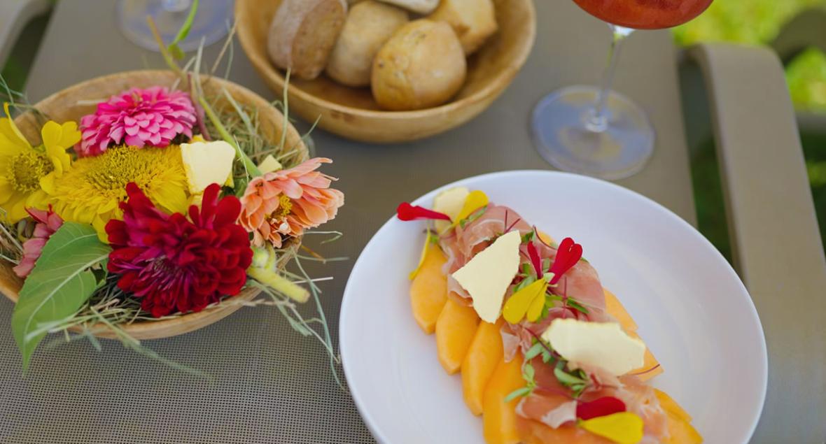 Tallerken med skinke og melon, brød, blomster og en cocktail.