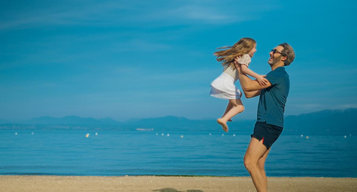 Vater und Tochter spielen fröhlich am Strand unter blauem Himmel.