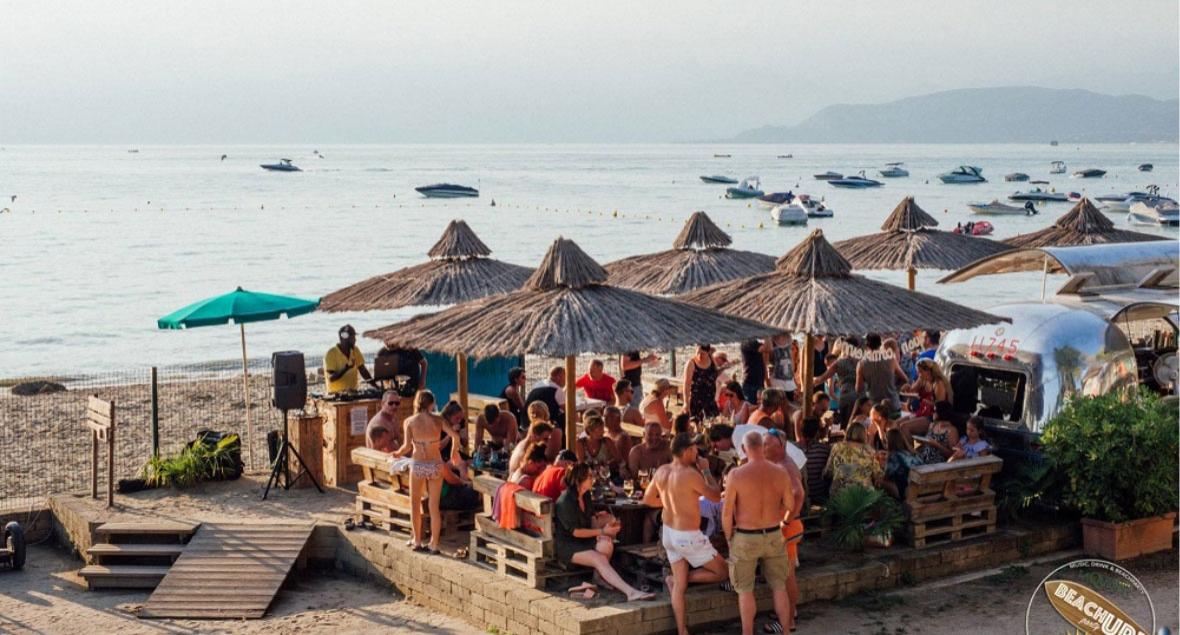 Crowded beach with umbrellas, bar, and boats on the sea.