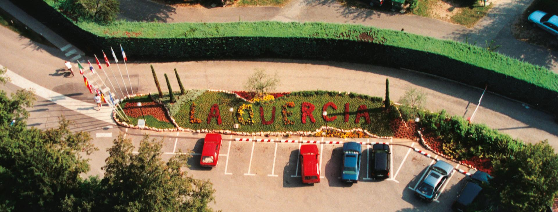 Indgang til La Quercia med blomsterbed og parkeringsplads.