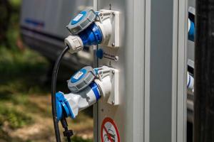 Elektrische Säule mit blauen Steckern, nahe einem Wohnmobil im sonnigen Campingplatz.