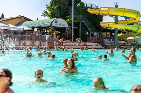 Piscina affollata con scivolo giallo e persone che si divertono.